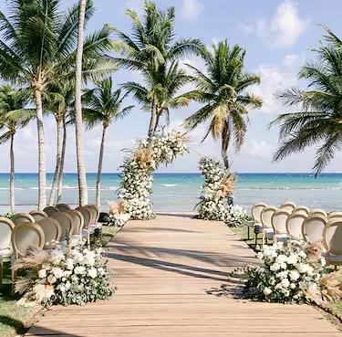 A wooden aisle between white chairs and floral arches on a palm-lined beach, set for a wedding ceremony