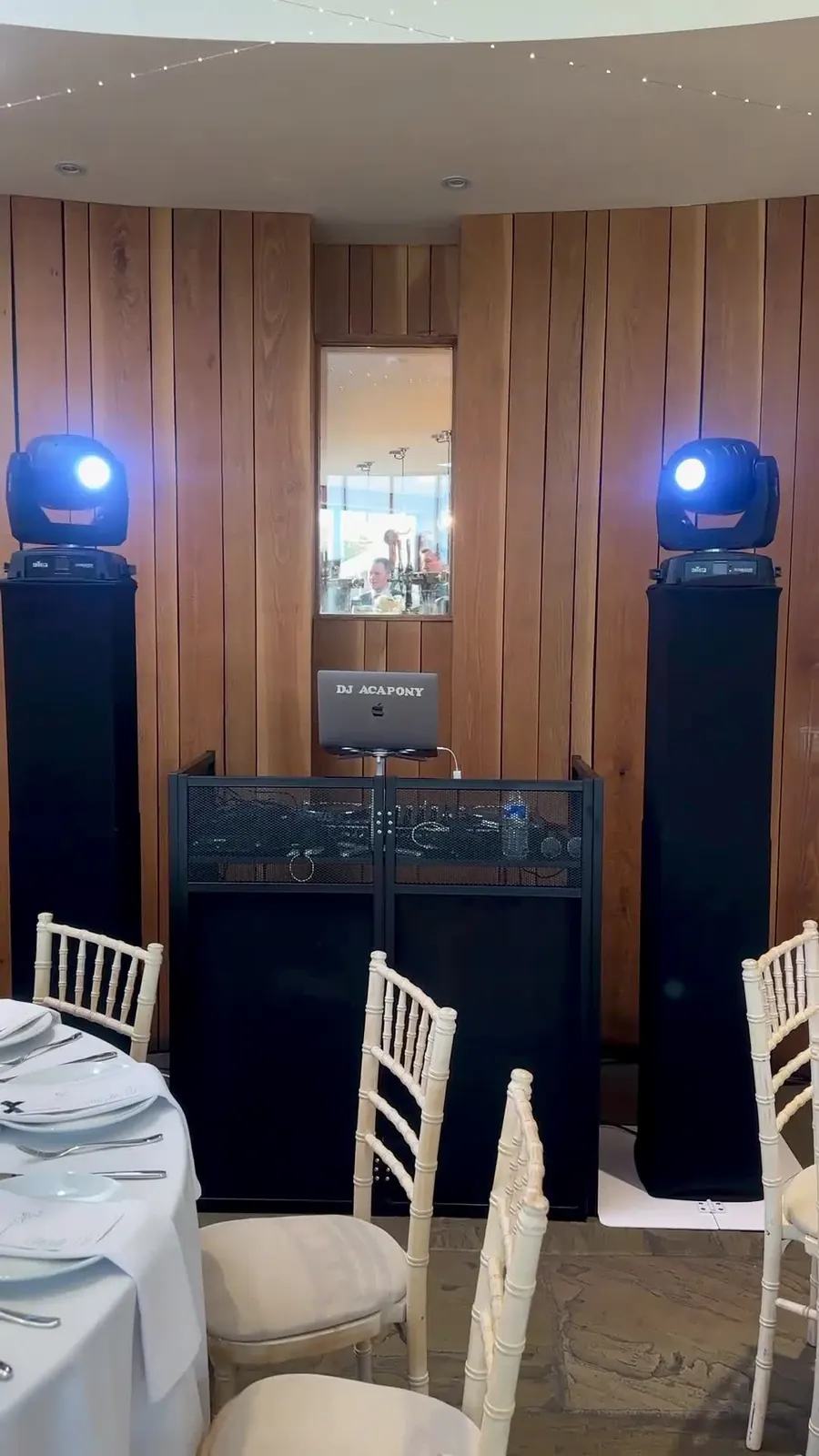 Two moving-head lights on black stands either side of a DJ booth at a wedding reception