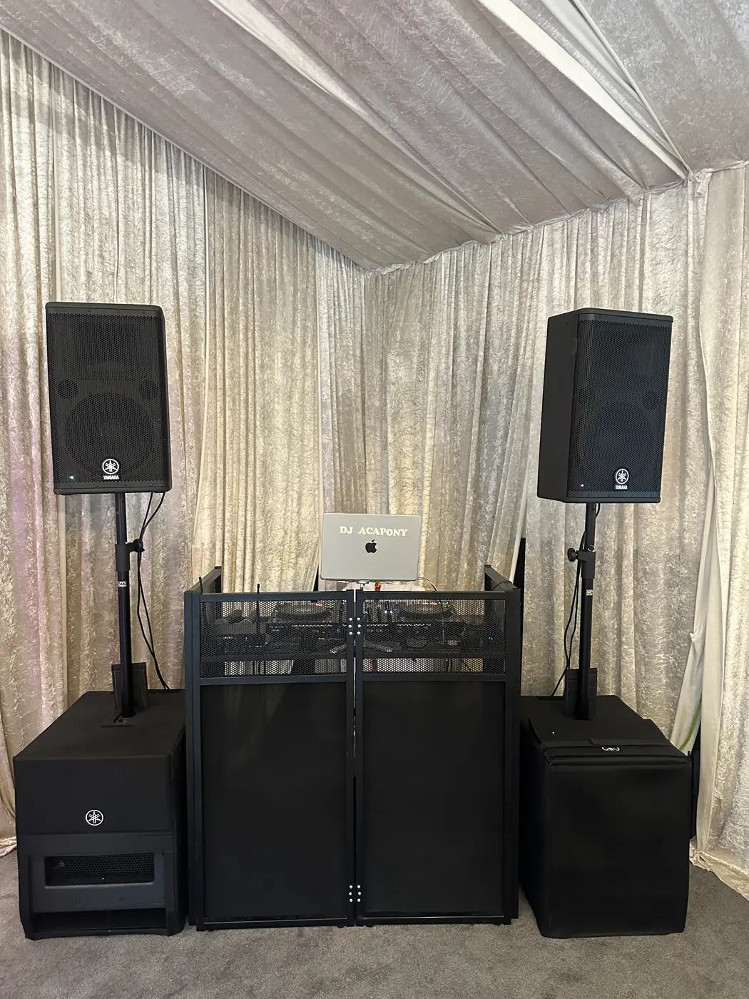 A black DJ booth with two speakers on stands and subwoofers, set up in a draped room