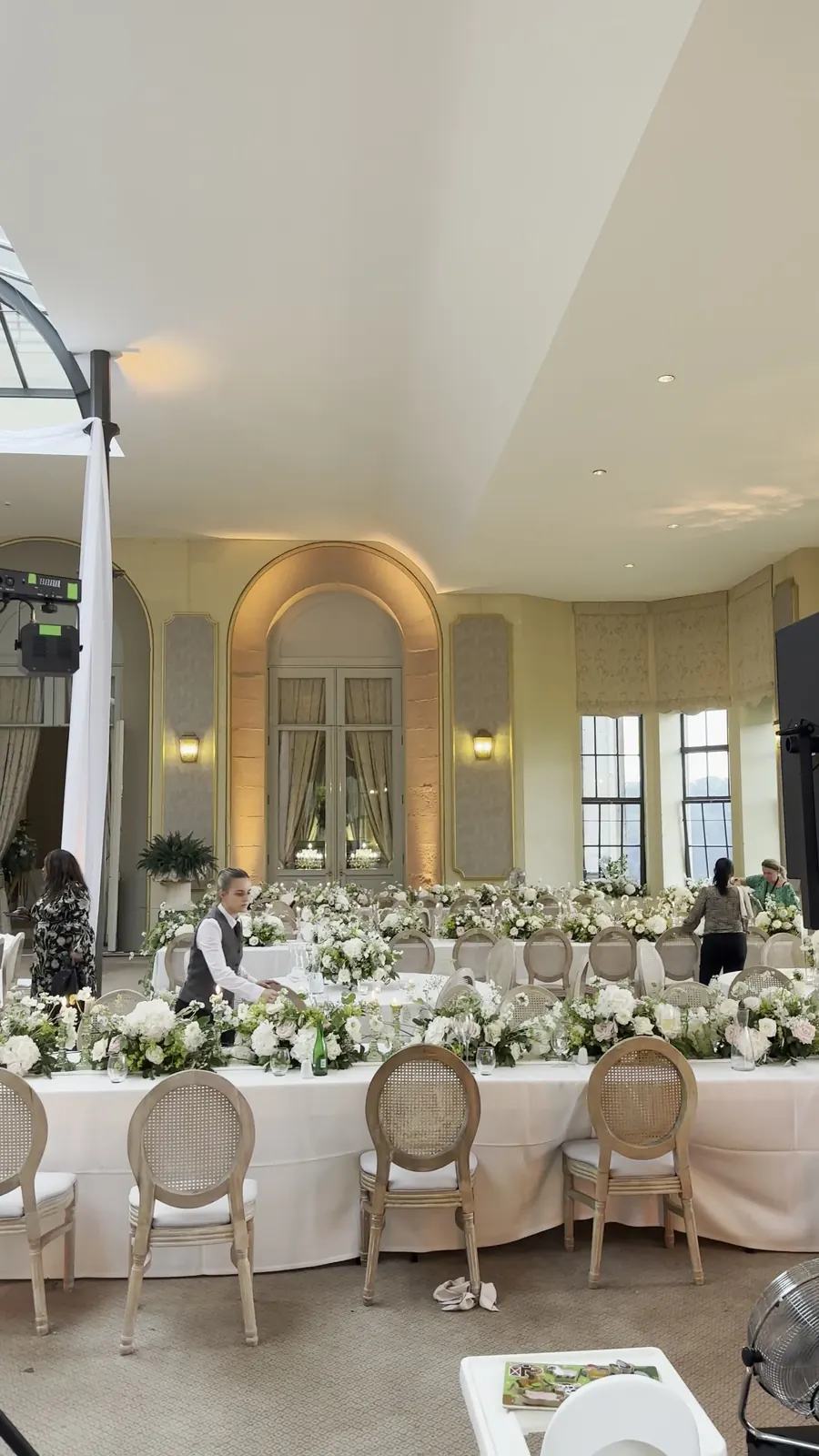 A venue being set for a dinner, with long tables of white flowers under arched windows