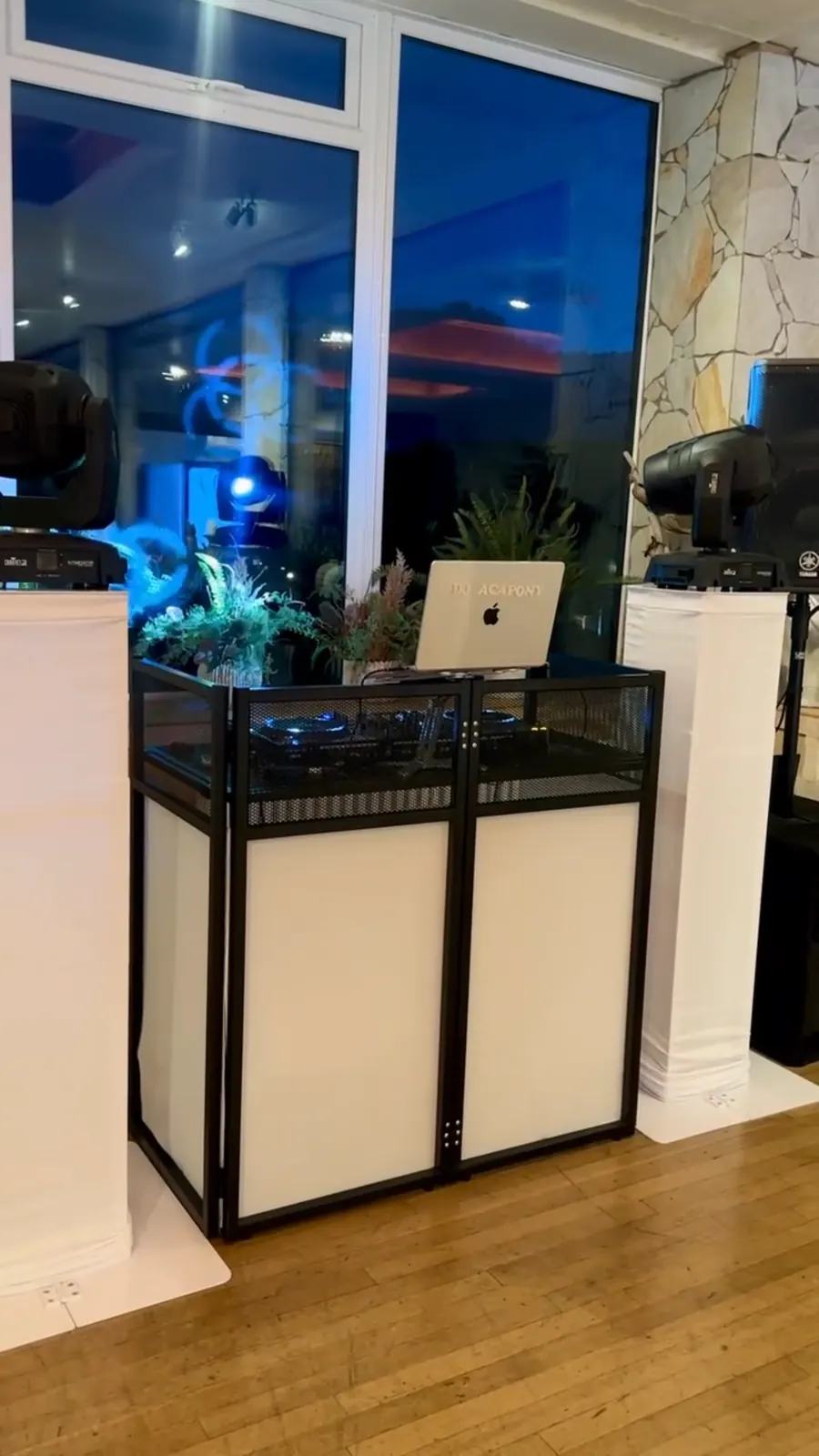 A white DJ booth with speakers and moving-head lights set up by a window at a venue